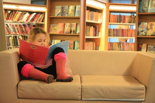A small girl is curled up on a couch reading a book in her local kids library.