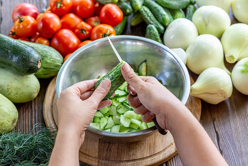 Someone roughly cutting small cucumbers into a metal bowl that is surrounded with various vegetables.