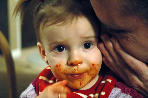 A baby girl with her face covered in red marinara sauce from eating spaghetti with her family.