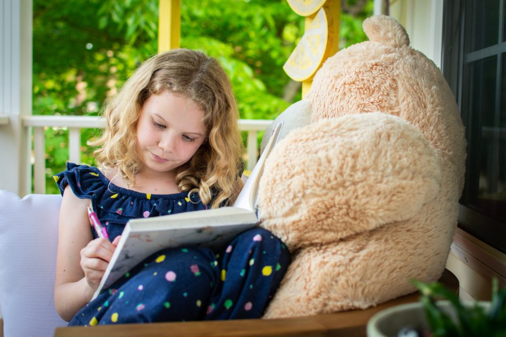 a girl in a blue dress sitting on a porch reading with a large stuffed bear.