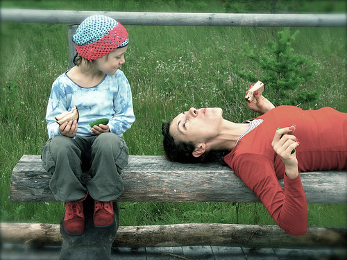 A mother and her daughter enjoying a snack while taking a break on a wooden park bench.