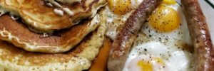 A diner plate with freshly made pancakes covered in butter and syrup alongside sausages and fried eggs.