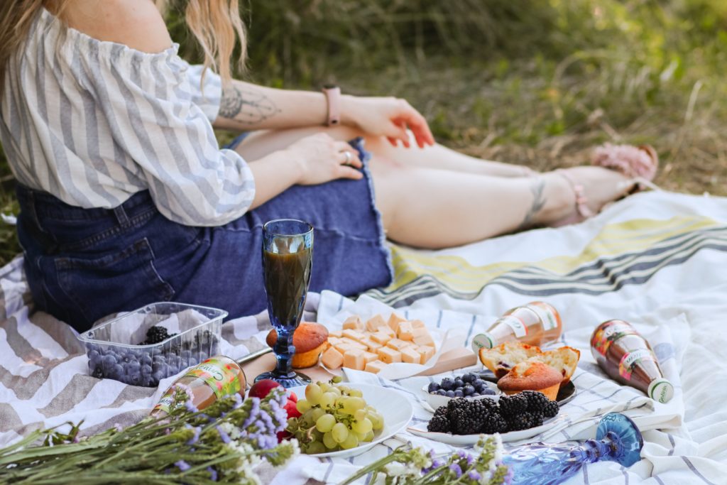 A woman in a striped top and denim skirt sitting on a blanket with a picnic at a local park.