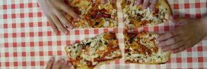 six hands reaching in to grab a piece of pizza on a red and white checkered tablecloth.