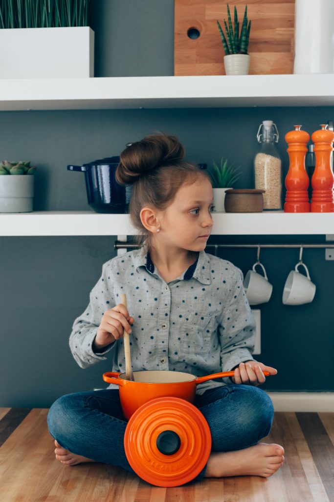 A young girl sitting on the kitchen counter stirring something in a small orang cast-iron pot with a wooden spoon.