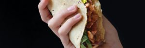A human hand holding a fesh chicken taco in a corn tortilla against a black background.