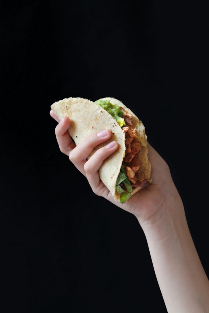 A human hand holding a fesh chicken taco in a corn tortilla against a black background.