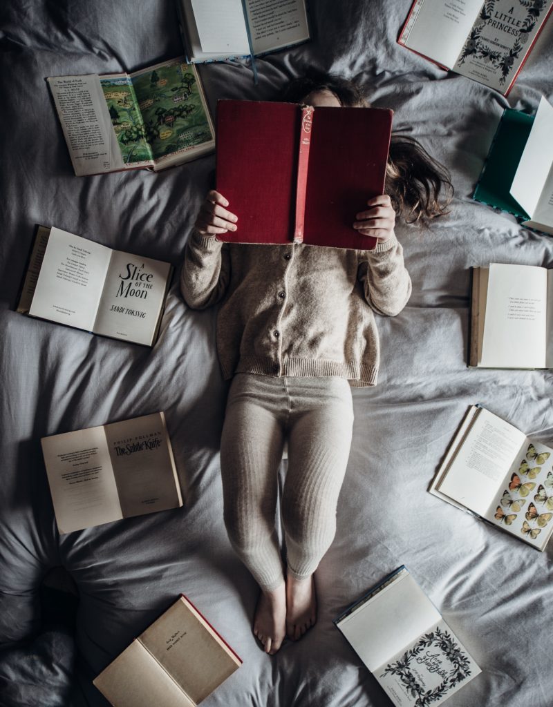 A small girl laying on a bed reading while surrounded by open books.
