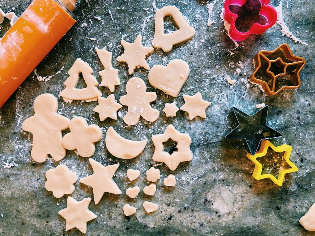 Sugar cookies being cut out into various shapes by cookie cutters on a flour dusted marble counter top.