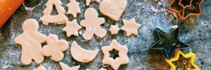 Sugar cookies being cut out into various shapes by cookie cutters on a flour dusted marble counter top.