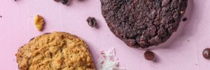 Several delicious cookies of various types on a pink table surrounded by walnuts raisins and chocolate chips.