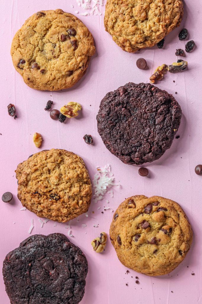 Several delicious cookies of various types on a pink table surrounded by walnuts raisins and chocolate chips.