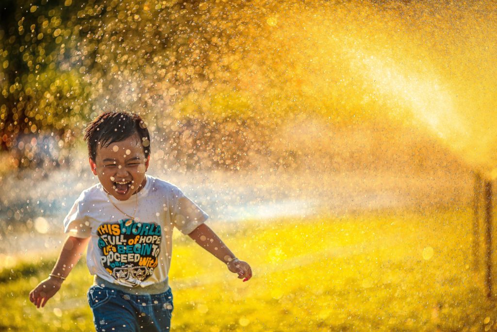 A young boy playing outside by running through the spray of a lawn sprinkler.