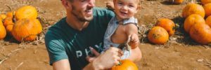 A man carries a smiling baby and a pumpkin during a day trip from The Woodlands, TX to a pumpkin patch