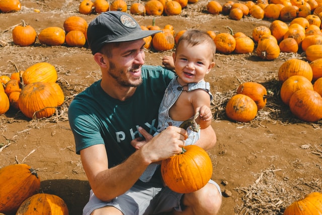 A man carries a smiling baby and a pumpkin during a day trip from The Woodlands, TX to a pumpkin patch