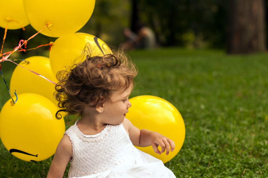 Little girl sitting next to yellow balloons.