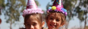 girl in left blowing purple balloon beside girl wearing pink birthday hat