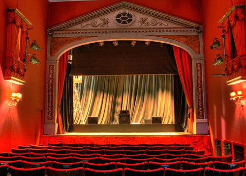 Image of a live theatre with red curtains and red seating.