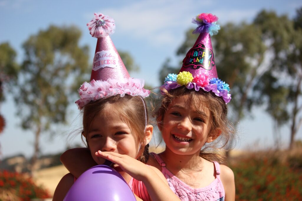 Two small girls in party hats blow up a balloon at a family-friendly venue in Leawood, KS