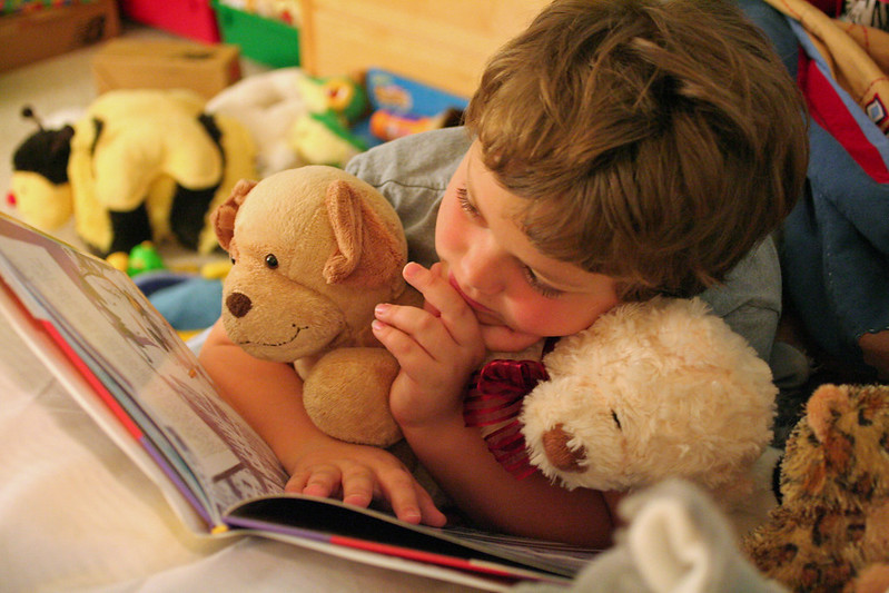 A child holding a stuffed animal reading a book.