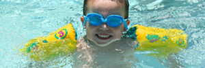 A smiling child in a pool with blue google and floaties on his arms.