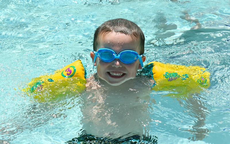 A smiling child in a pool with blue google and floaties on his arms.