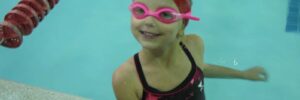 A little girl in a pool during a swim class smiling with pink goggles and a red swim cap