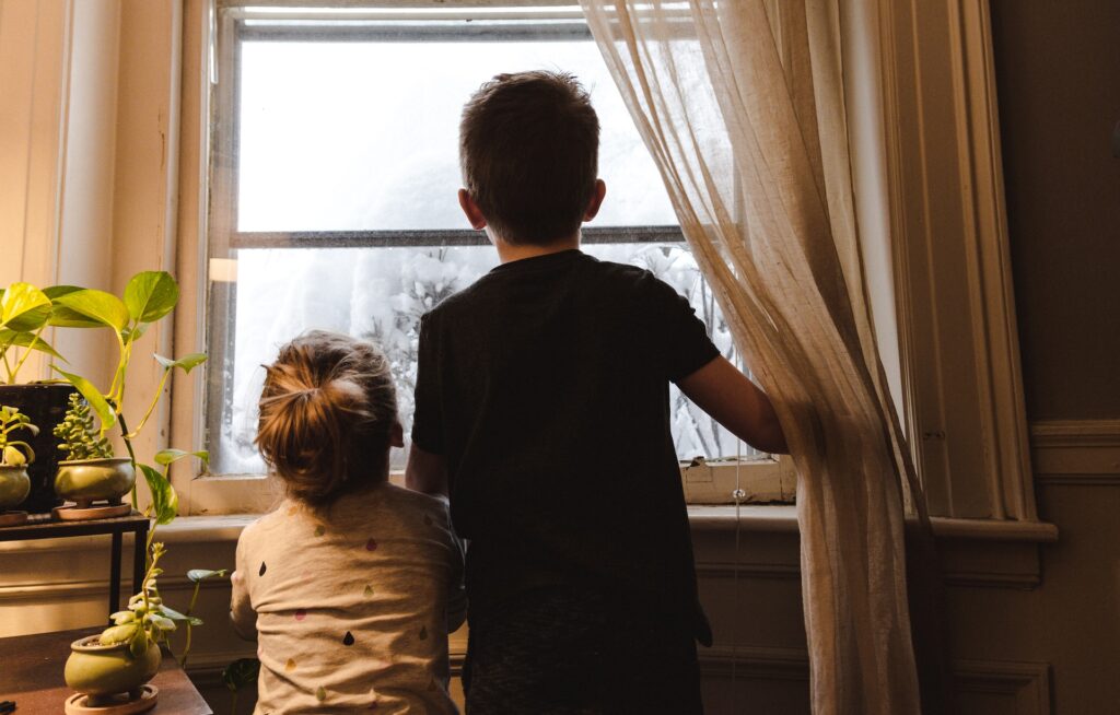 A young boy and a little girl looking outside their window seeing all the snow and frost.