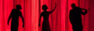 Three black silhouettes behind a red curtain on a stage.