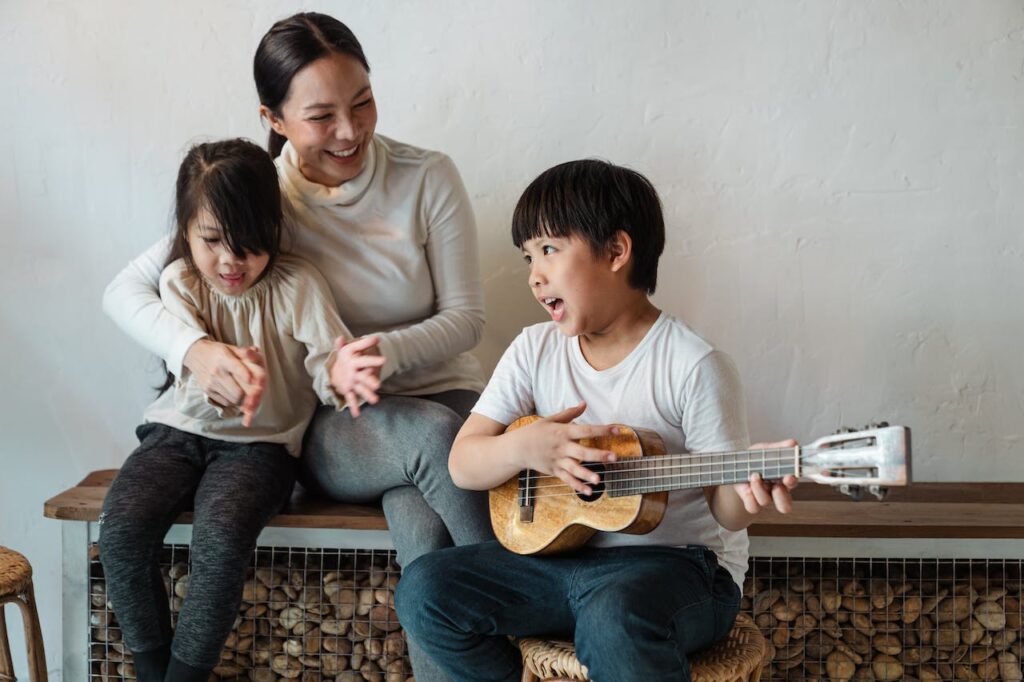 Young child playing guitar and singing for his mother and another young child.