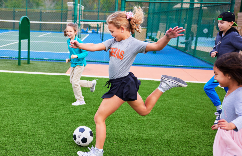 A young girl in a Texas shirt kicking a soccer ball on a field during an outdoor summer camp game with other kids.