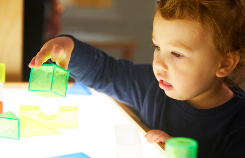 A young toddler in a blue sweater, playing with clear green and yellow blocks during an early childhood education class.