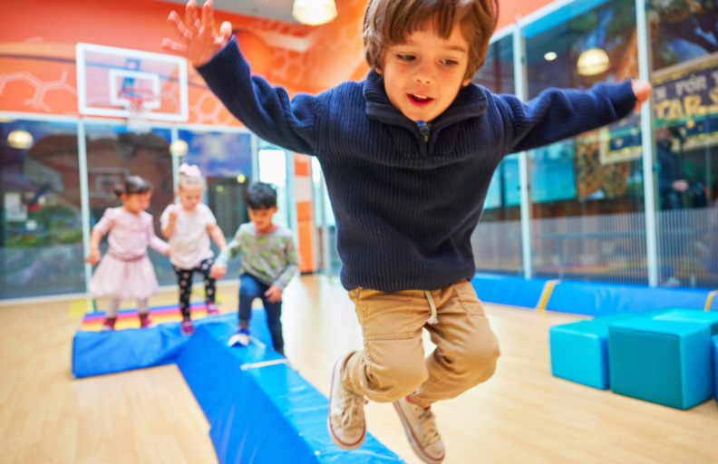 A toddler in a sweater jumping off of a blue mat with 3 other children following behind at a Crème de la Crème daycare school.