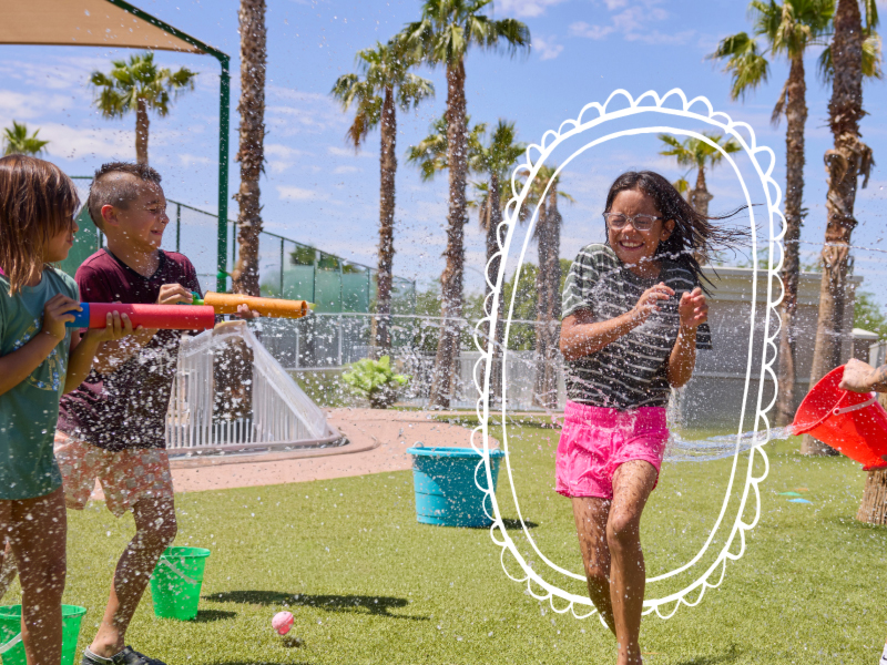 A young girl in a grey shirt kicking a soccer ball on a field during an outdoor summer camp game with other kids.