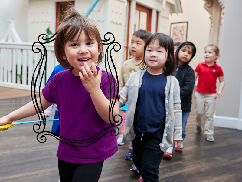 A young girl in a grey shirt kicking a soccer ball on a field during an outdoor summer camp game with other kids.